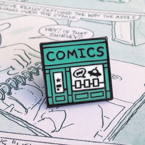 以下が含まれることがあります： コミックストアの形をしたターコイズブルーのエナメルピンで、店先に「COMICS」の文字があります。店先には、吹き出し、UFO、コミックが3冊入ったショーウィンドウがあります。ドアには星と線が描かれています。