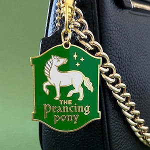Puede incluir: Un charm de metal dorado con fondo verde e ilustración de caballo blanco. El charm dice "The Prancing Pony" en letras doradas y cuelga de una cadena dorada sujeta a un bolso negro.