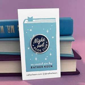 Puede incluir: Un pin de esmalte circular con fondo azul oscuro y texto plateado que dice Night owl BOOK CLUB. El pin está sujeto a una tarjeta de presentación blanca con una ilustración de un libro azul claro con estrellas blancas y el texto RATHER KEEN.