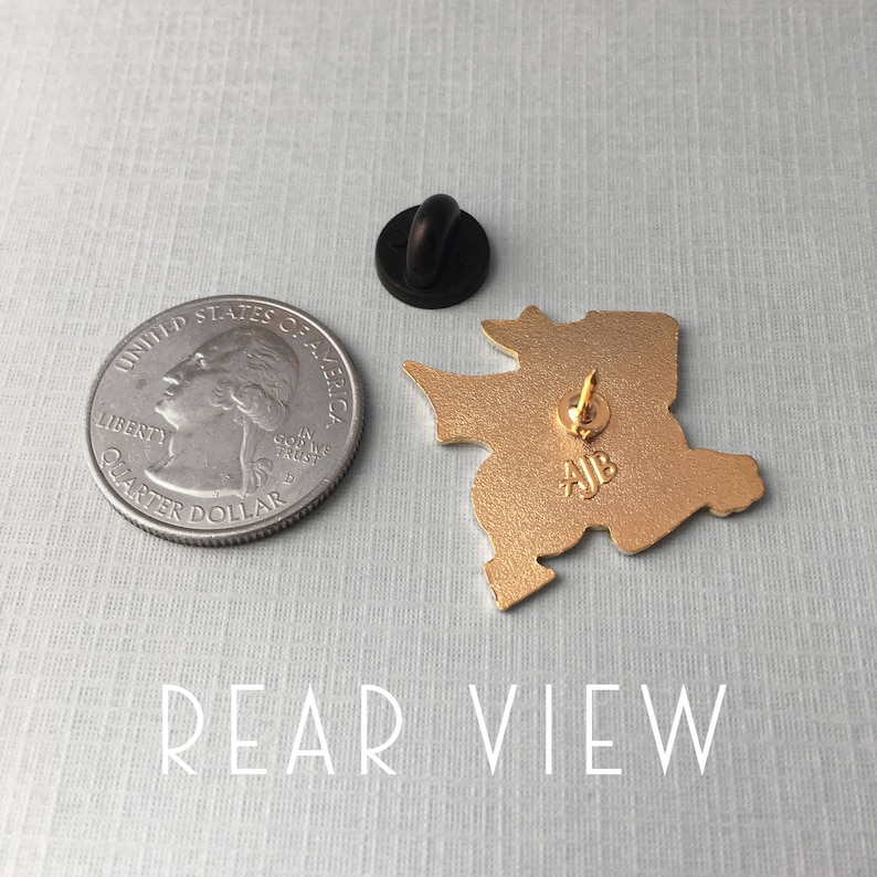 May include: Gold-colored enamel pin featuring a cartoon-style character with a large head and a small body. The pin has a black rubber clutch back and the text "AJB" is visible on the back.