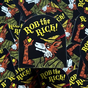 May include: Black sticker featuring a cartoon fox in a Robin Hood-style hat, holding a golden orb. The text "ROB the RICH!" is in yellow, arched above the fox. The fox is in a green and gold outfit.
