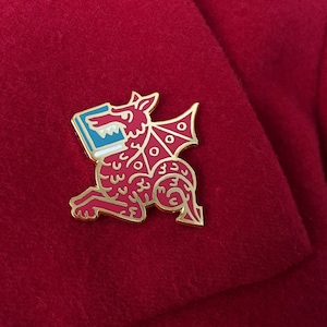 May include: Gold-colored enamel pin of a red dragon holding a book. The dragon has a detailed design with white and blue accents.