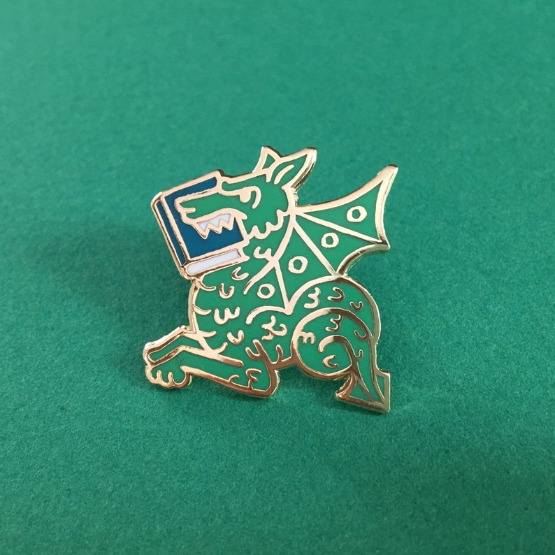 May include: A green and gold enamel pin of a dragon holding a book in its mouth. The dragon has a white and blue book in its mouth and a gold outline.