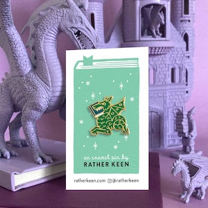 May include: A green enamel pin of a dragon holding a book. The pin is on a light green background with white stars. The text on the card reads "an enamel pin by RATHER KEEN ratherkeen.com @ratherkeen"