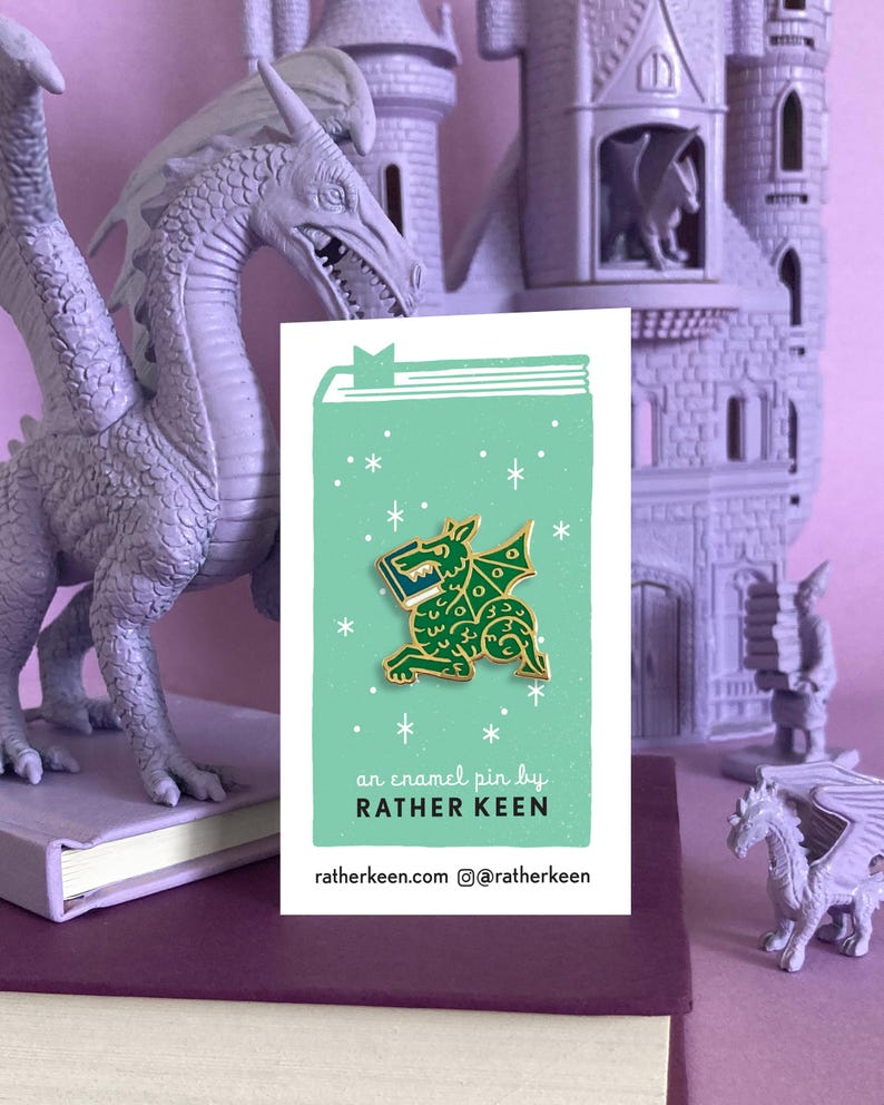 May include: A green enamel pin of a dragon holding a book. The pin is on a light green background with white stars. The text on the card reads "an enamel pin by RATHER KEEN ratherkeen.com @ratherkeen"