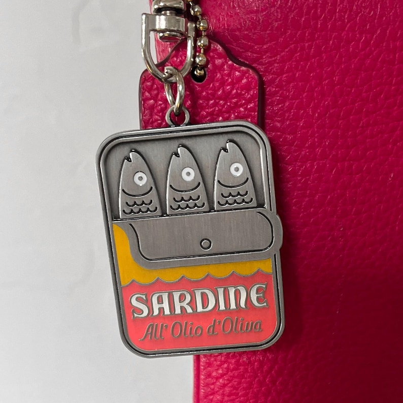 May include: A silver-colored sardine can charm with three fish illustrations, yellow and red accents, and the word "SARDINE" in red. The charm hangs from a silver chain and is set against a pink leather background.