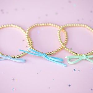 May include: Three gold beaded bracelets with delicate bows in shades of blue and green. The bracelets are arranged on a pink background with scattered glitter.