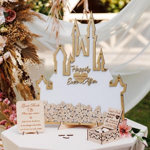 May include: A castle-shaped wooden guest book frame with a white center, featuring the text "Happily Ever After" and a heart with "Samuel & Dakota 09.02.2027". Includes a small wooden sign, a box of heart-shaped tokens, and many signed hearts inside.