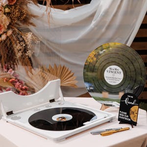 May include: A white and black decorative record player with a black vinyl record. A large, round, reflective disc with the names "Mr & Mrs Vincent" and a date is displayed. A small sign reads "For the Record, Please Sign Our Guestbook."