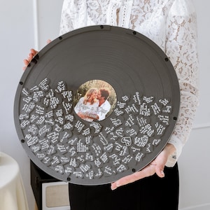 May include: A large, grey vinyl record-shaped guest book with a photo in the centre. Small, heart-shaped pieces surround the photo, each with handwritten messages. The record is held by a person wearing a white lace top.