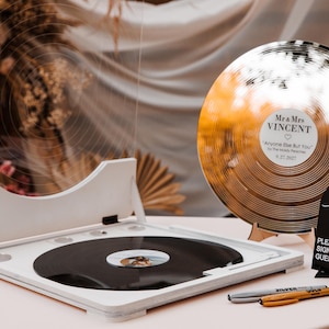 May include: A white wooden record player with a black vinyl record, a gold-coloured vinyl record with the names "Mr & Mrs VINCENT" and a sign that says "For the Record". Two pens are on the table. The theme is weddings.