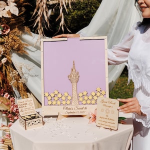 May include: A wooden guest book sign with a lavender background and a laser-cut tower design. The sign includes the text "Olivia's Sweet 16 September 13, 2027" and is accompanied by a small box and a sign that says "Guest Book".
