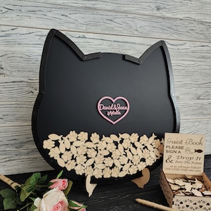 May include: A black cat-shaped guest book with a pink heart-shaped accent that reads "David & Jenn 3/9/2024." The guest book has wooden paw prints and fish cutouts. A sign reads "Guest Book, Please Sign A or Drop It Into The Frame."