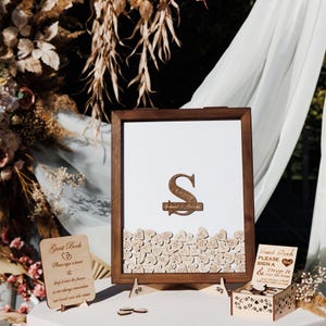 May include: A wooden framed guest book with a white background and a wooden initial "S" and the text "Est 2017". The frame is filled with small wooden hearts. Additional wooden signs and a box are also present.
