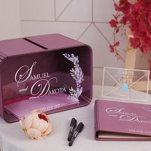 May include: A plum-colored wedding card box, guest book, and sign. The box and book are inscribed with "Samuel and Dakota" and the date "09.02.27". Floral accents and a clear acrylic sign that reads "Cards and Gifts" are also present.