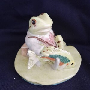 May include: A ceramic figurine of a frog wearing a suit and holding a fish. The frog is sitting on a lily pad.