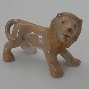 May include: A small, glossy, light brown ceramic lion figurine standing in a profile position. The lion features a sculpted mane, a curved tail, and a slightly open mouth with visible teeth. The surface has a smooth, reflective finish.