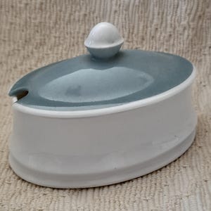 May include: A white ceramic oval-shaped dish with a light blue lid and a small spout. The lid has a white knob on top.