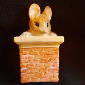 May include: A ceramic figurine of a brown mouse peeking out of a brick chimney. The mouse has large ears and black eyes. The chimney is made of orange and white bricks.