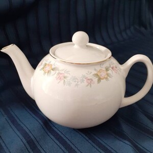 May include: A white teapot with a floral design in shades of pink, yellow, and green. The teapot has a gold trim around the lid and a gold band around the body.