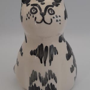 May include: A ceramic cat figurine with a glossy off-white finish and hand-painted black markings. The cat features a stylised face with simple black line details for the eyes, nose, and mouth, and scattered black brushstroke spots across its body.