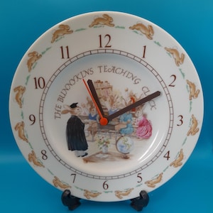 May include: A white plate clock with a bunny theme. The clock face has black numerals, brown clock hands, and the text "THE BUNNYKINS TEACHING CLOCK". The clock is supported by a black stand and has a blue background.