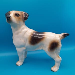 May include: A ceramic Jack Russell Terrier dog figurine. The dog is white with brown and black patches. It is standing with its head raised, and its tail is slightly up. The figurine has a glossy finish and is set against a blue background.