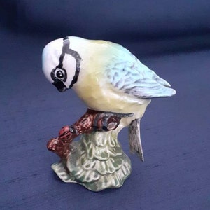 May include: A bluebird figurine perched on a branch with green leaves. The bird is painted in shades of blue, yellow, and white, with black eyes and a black beak. The figurine is made of ceramic and is approximately 4 inches tall.