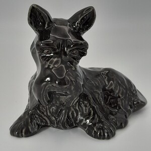 May include: A glossy black ceramic Scottish Terrier dog figurine in a resting pose. The dog has detailed fur texture, upright ears, and a distinctive beard. A decorative item.