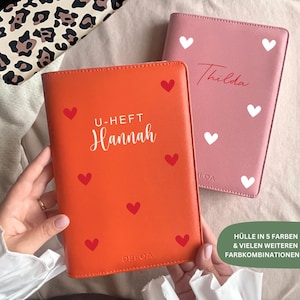 Puede incluir: Dos cubiertas de polipiel, una naranja y otra rosa, decoradas con corazones. La cubierta naranja tiene el nombre "Hannah" y el texto "U-HEFT" impresos en blanco. La cubierta rosa tiene el nombre "Thilda" impreso en rojo. Las cubiertas están decoradas con corazones rojos y blancos.
