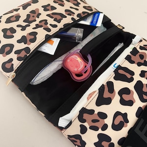 Puede incluir: Una bolsa de pa&ntilde;ales de estampado de leopardo con una cremallera negra y un asa negra. La bolsa est&aacute; abierta y llena de art&iacute;culos para beb&eacute;s, incluyendo pa&ntilde;ales, toallitas, un chupete y desinfectante de manos.