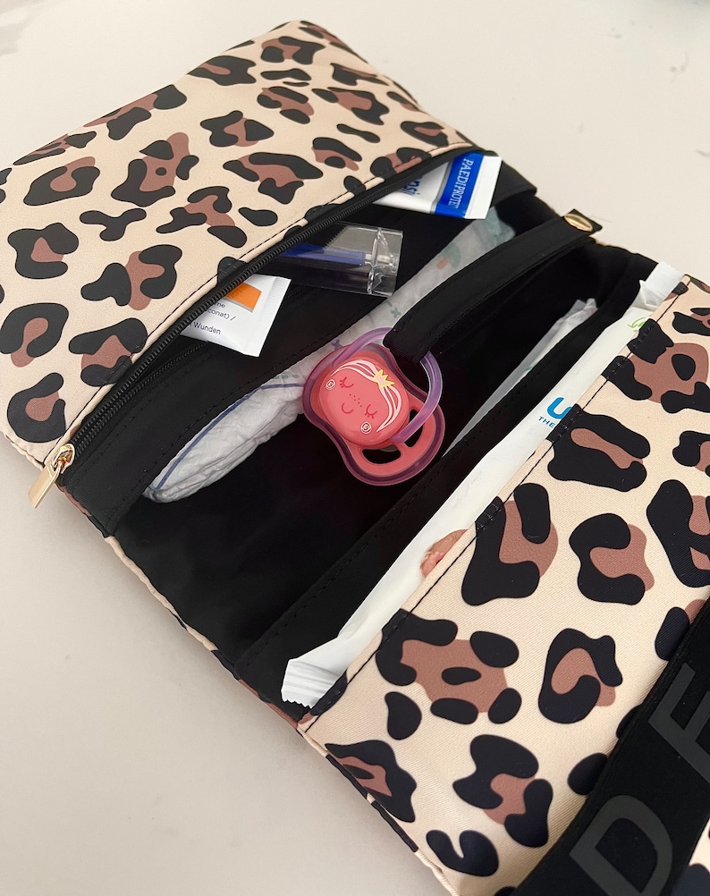 Puede incluir: Una bolsa de pa&ntilde;ales de estampado de leopardo con una cremallera negra y un asa negra. La bolsa est&aacute; abierta y llena de art&iacute;culos para beb&eacute;s, incluyendo pa&ntilde;ales, toallitas, un chupete y desinfectante de manos.