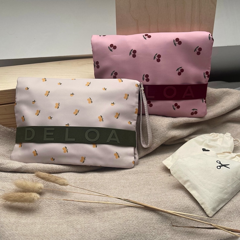 Puede incluir: Dos bolsas de tela rosa y beige con estampado floral y las palabras "DELOA" y "LOA" impresas en ellas. Las bolsas est&aacute;n sobre una superficie de tela beige.