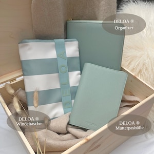 Puede incluir: Una caja de madera llena de tres bolsas de tela a rayas blancas y azul claro. Las bolsas est&aacute;n etiquetadas con "DELOA&reg;" y tienen diferentes usos: "Organizador", "Windeltasche" y "Mutterpasch&uuml;lle".