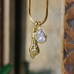 May include: A gold chain necklace with a small hand charm and a teardrop-shaped crystal pendant.