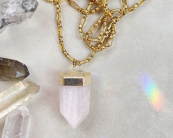 May include: A gold chain necklace with a rose quartz pendant. The pendant is a point with a gold cap at the top.