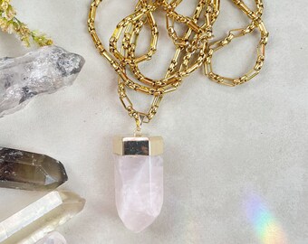 May include: A gold chain necklace with a rose quartz pendant. The pendant is a long, pointed crystal with a gold cap at the top. The necklace is lying on a white surface with other crystals.