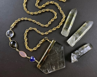 May include: A gold chain necklace with a large, clear quartz pendant. The pendant is shaped like a triangle and has a gold trim. The necklace also features several smaller gemstone beads in shades of blue, pink, and brown.