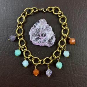 May include: A gold chain bracelet with multiple charms. The charms are different colored gemstones, including purple, green, and orange. The bracelet is laying on a brown surface with a large pink crystal.