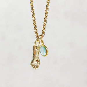 Puede incluir: Collar de cadena dorada con un dije de puño y un colgante de piedra preciosa azul claro.