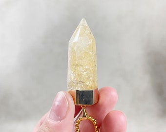 May include: A gold-toned pendant featuring a clear, yellow crystal point with a gold-toned setting. The pendant is attached to a gold-toned chain.