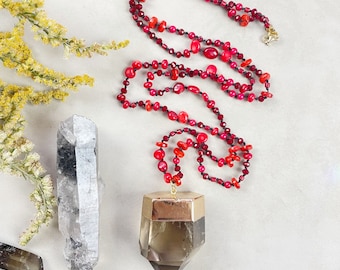 May include: A long red and black beaded necklace with a large, faceted, clear quartz pendant. The pendant is set in a gold-toned metal frame.