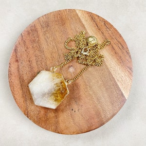 Peut inclure: Un collier en chaîne dorée avec un grand pendentif hexagonal en citrine clair et jaune. Le pendentif est sur une surface en bois.