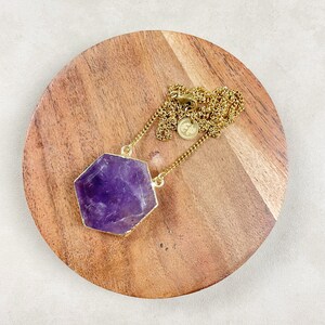 Peut inclure: Un collier en chaîne dorée avec un grand pendentif en améthyste facetté. Le pendentif est de forme hexagonale et a une couleur violet foncé.