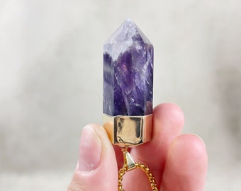 May include: A purple amethyst crystal point pendant with a gold-toned chain and a gold-toned setting. The pendant is held in a hand.