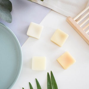 Puede incluir: Cuatro pequeñas barras de jabón cuadradas, dos blancas y dos blanquecinas, sobre una superficie blanca con una rama de hojas verdes. Las barras de jabón están dispuestas en forma de diamante.