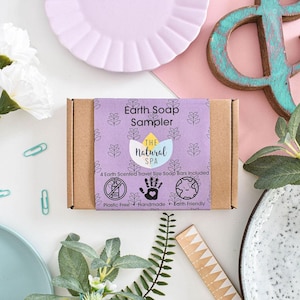 Könnte beinhalten: Eine braune Box mit einem lila und weißen Etikett, auf dem "Earth Soap Sampler" und "The Natural Spa" steht. Das Etikett enthält auch Symbole für plastikfrei, handgefertigt und umweltfreundlich. Die Box enthält 4 erdschnuppernde Reise-Seifenstücke.