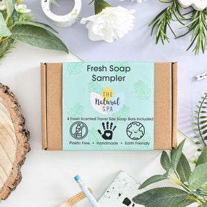 Könnte beinhalten: Eine Schachtel mit natürlichen Seifenstücken mit dem Text "Fresh Soap Sampler" und "The Natural Spa" auf der Vorderseite. Die Schachtel ist mit grünen Blättern und dem Text "4 Fresh Scented Travel Size Soap Bars Included" verziert. Die Schachtel enthält auch den Text "Plastic Free", "Handmade" und "Earth Friendly".