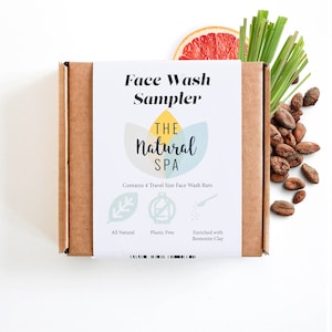 Può includere: Una scatola di cartone marrone con un'etichetta bianca che dice "Face Wash Sampler" e "THE Natural SPA". L'etichetta include anche una grafica di un fiore con le parole "All Natural", "Plastic Free" e "Enriched with Bentonite Clay". La scatola è circondata da una fetta di pompelmo, erba verde e chicchi di cacao.