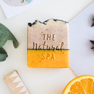 Op de afbeelding: Een rechthoekig stuk zeep met de tekst "THE Natural SPA" in reliëf. De bovenste helft is beige met donkere randen, de onderste helft is fel oranje. Een sinaasappelschijf en een logo zijn ook zichtbaar.